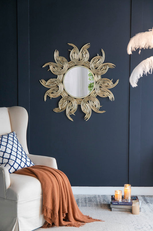 A Sunburst Metal Decorative Mirror in gold hangs on a dark blue wall above a cream armchair with a blue-and-white pillow and orange throw. Lit candles and stacked books are on the floor, with white feathers at the image's edge.