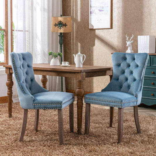 Two Nikki modern velvet tufted dining chairs with solid wood legs and nailhead trim are placed by a wooden table on a beige carpet in a cozy, well-lit room with framed art, a lamp, and decor in the background.
