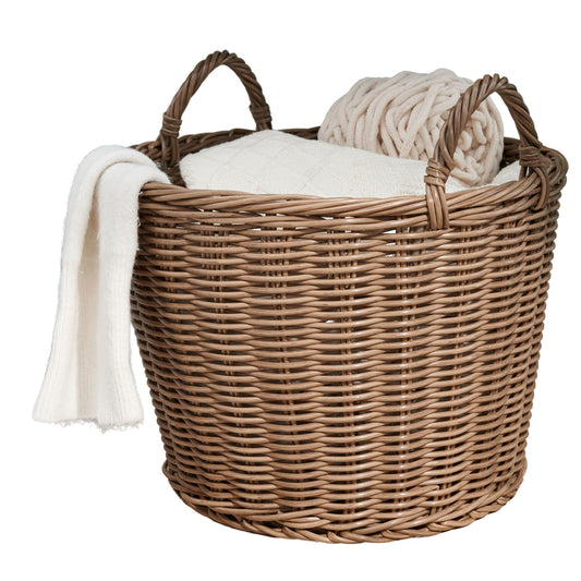 A Round Cone Woven Resin Wicker Basket with Handles in Walnut Brown holds a folded white blanket, a cream cushion, and a chunky knit throw draped over the edge.