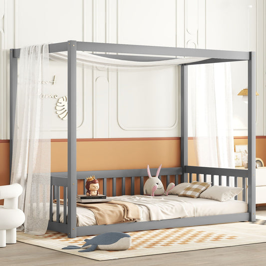 A cozy kids’ room features the Canopy Frame Floor Bed With Fence, Guardrails in gray, draped with white sheer fabric, topped with stuffed animals, a checkered pillow, beige blanket, toy crown; white walls have orange accents and decor.