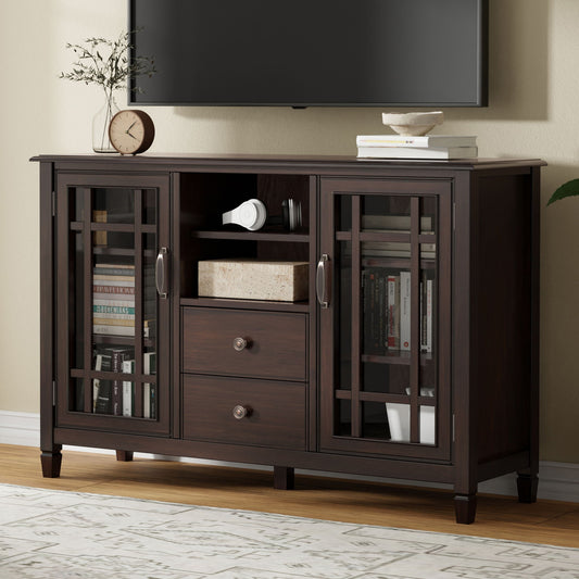 The Connaught Tall TV Stand in Brown features glass-paneled doors, two center drawers, and an open shelf. Books, decor, and electronics are neatly arranged inside and on top. A TV is wall-mounted above it against a beige background.