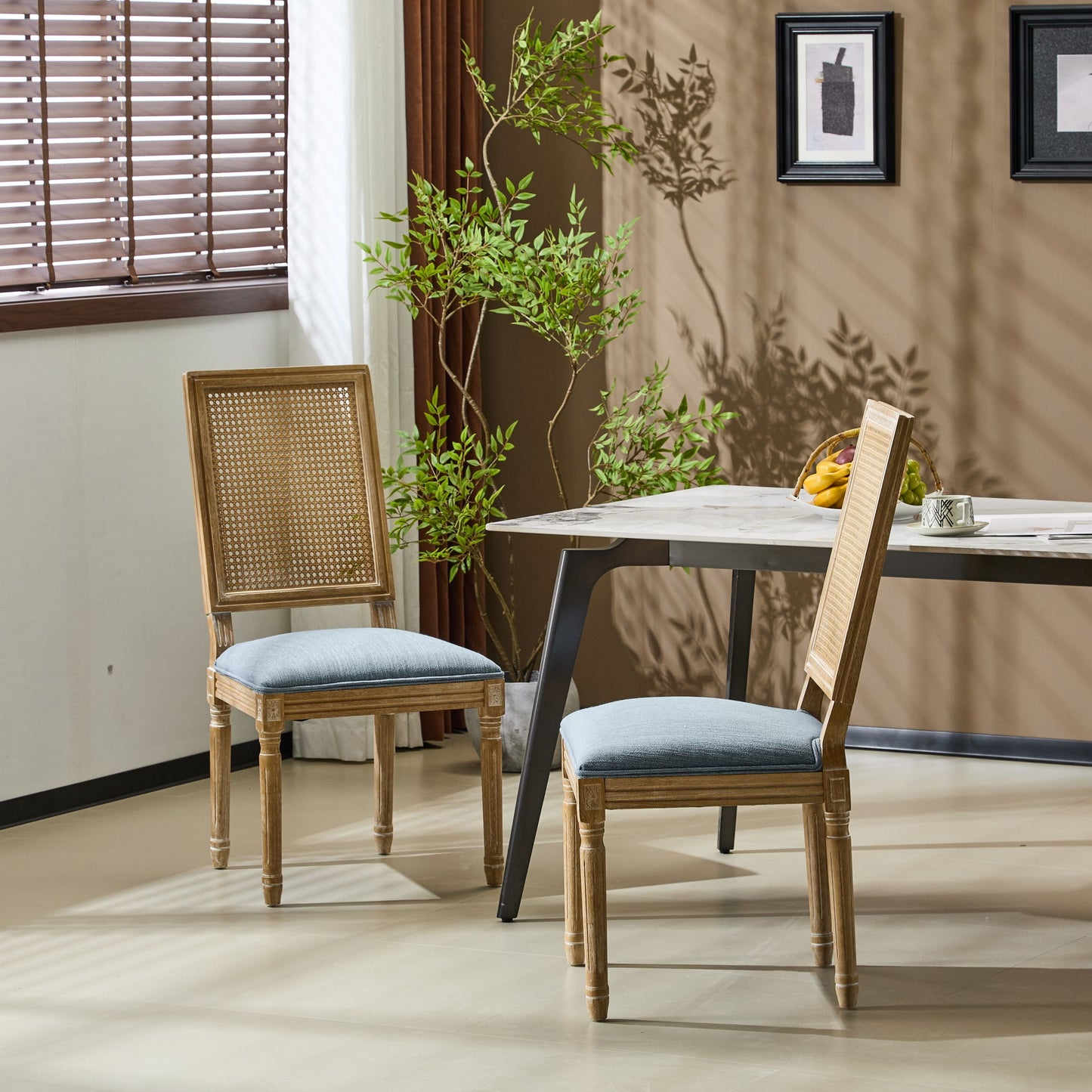 Two French Country Dining Chairs with rectangular cane backrests and smooth blue upholstered seats sit by a modern table in a bright room with beige walls, a potted plant, window blinds, and framed wall art.
