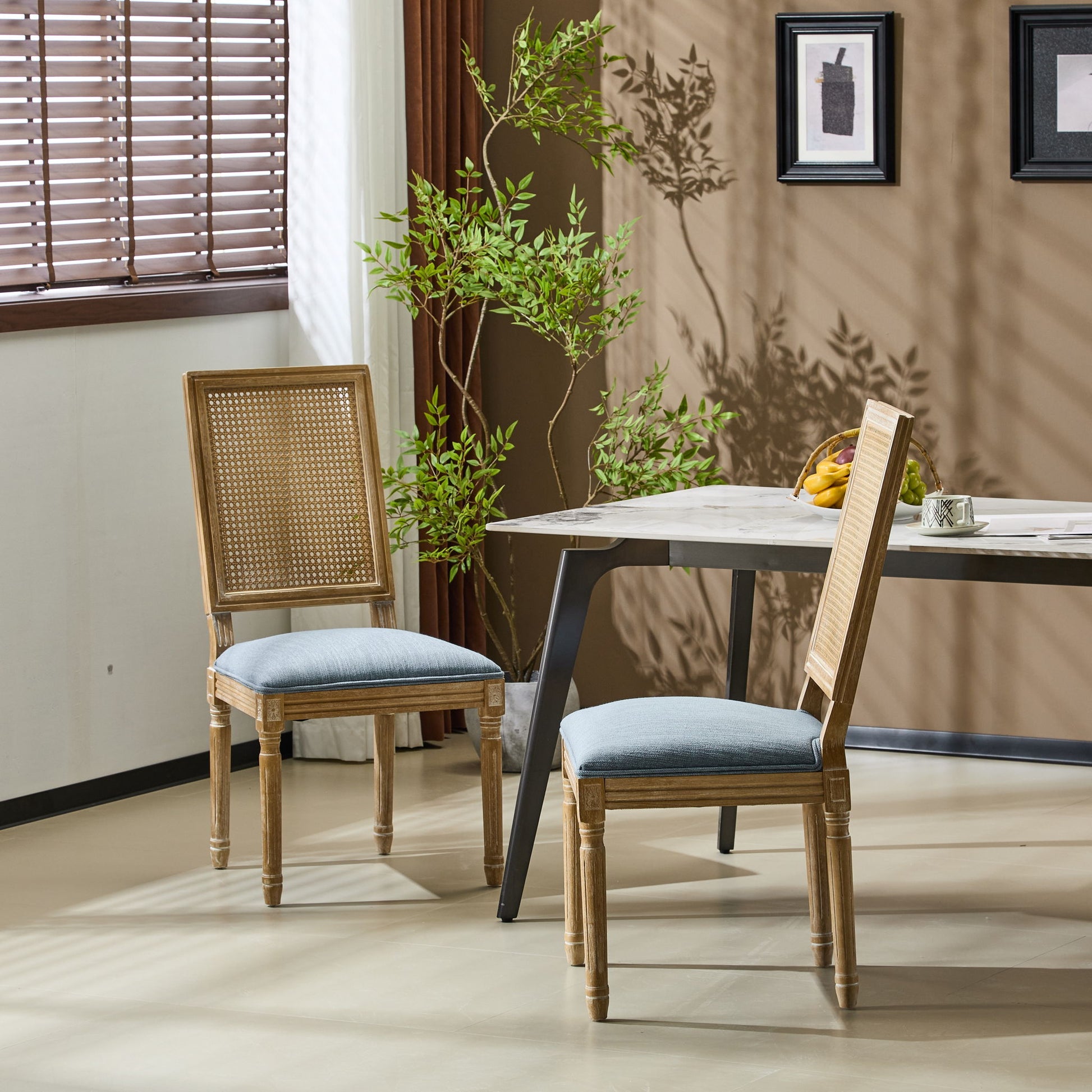 Two French Country Dining Chairs with rectangular cane backrests and smooth blue upholstered seats sit by a modern table in a bright room with beige walls, a potted plant, window blinds, and framed wall art.