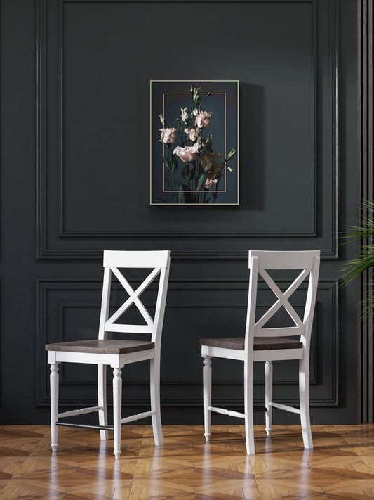 Two Mountina 24" Tall Bar Stools (Set of 2, Brown/White) with X-back design stand before a dark paneled wall. Above the stools is a framed floral painting, while a green plant peeks in from the right.