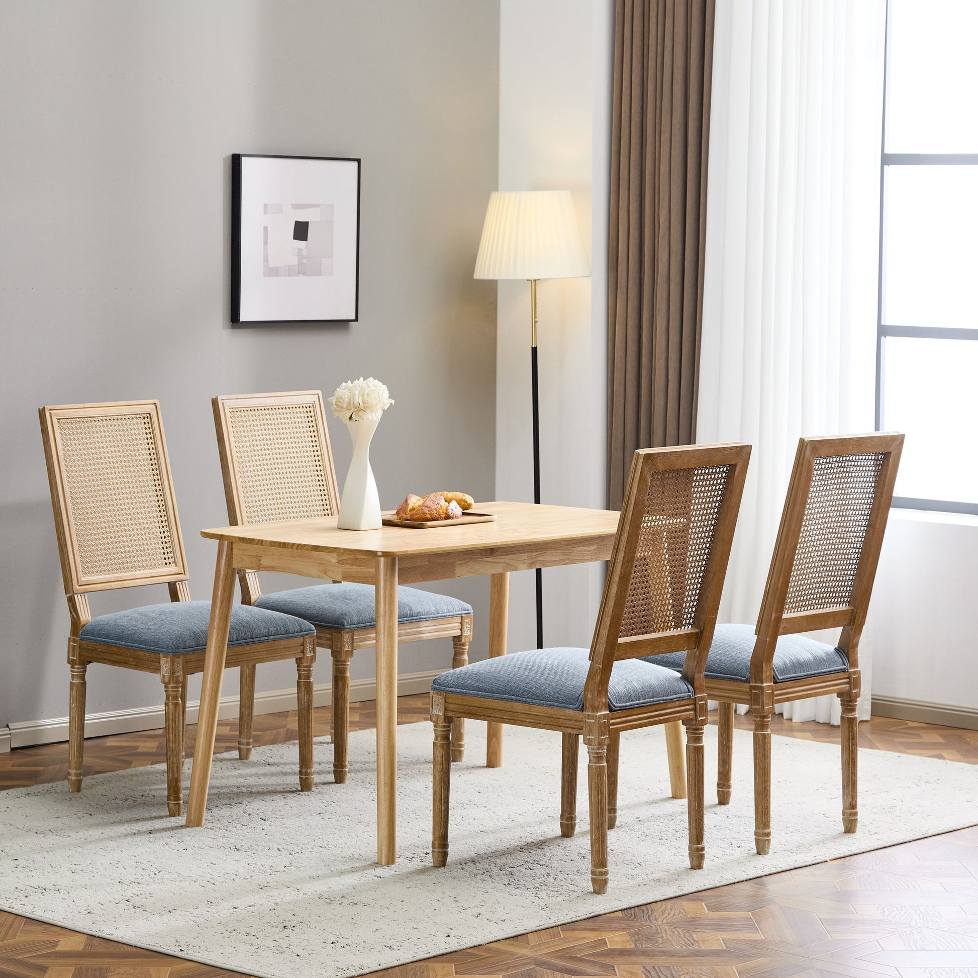 A modern dining room features a wooden table, four French Country Dining Chairs with rectangular cane backrests and smooth upholstered seating, a vase of white flowers, pastries on a plate, wall art, large curtained windows, and a floor lamp.
