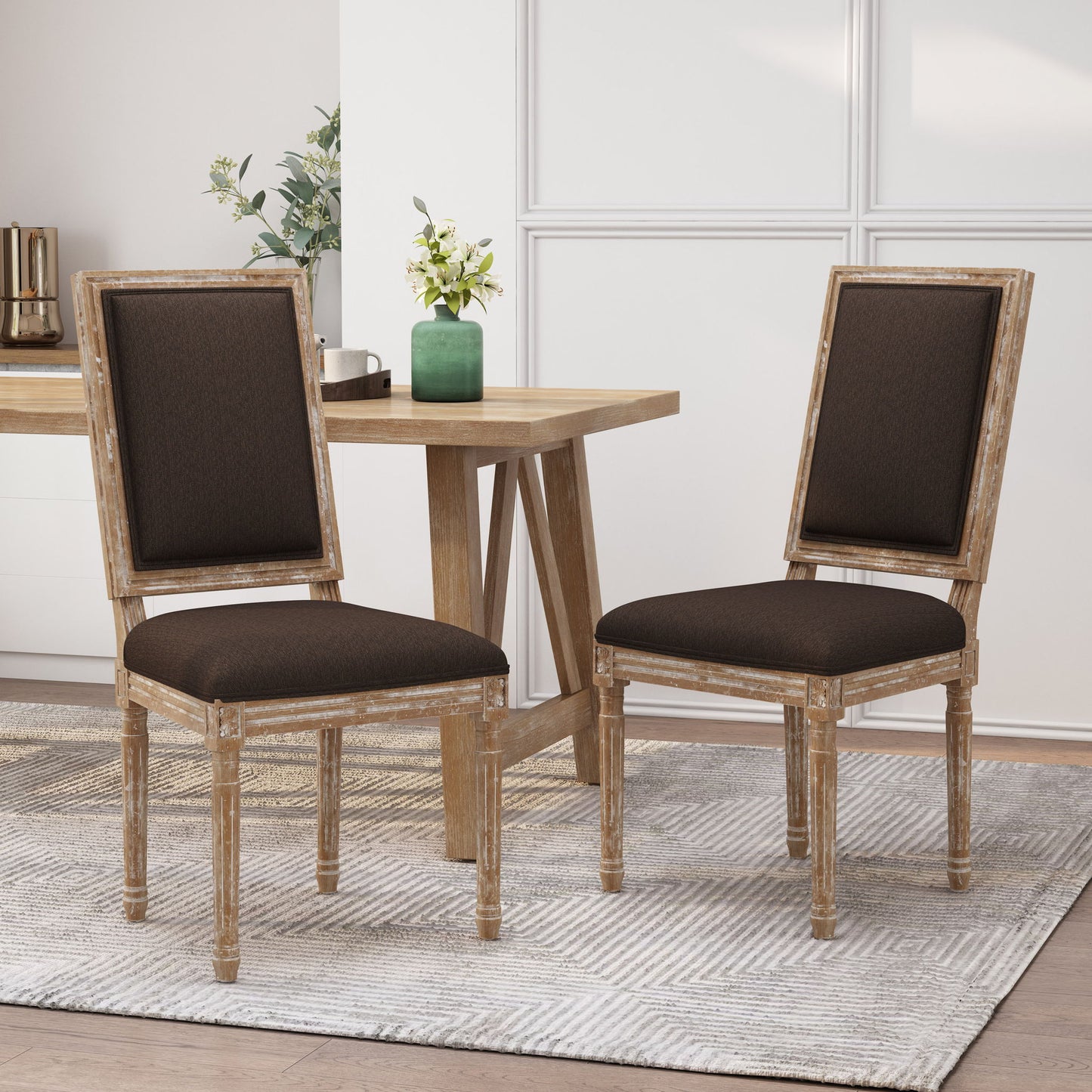 Two French Country Style Dining Chairs with upholstered seats and fluted legs sit on a light patterned rug next to a wooden table. A green vase with flowers and other decor can be seen in the background.