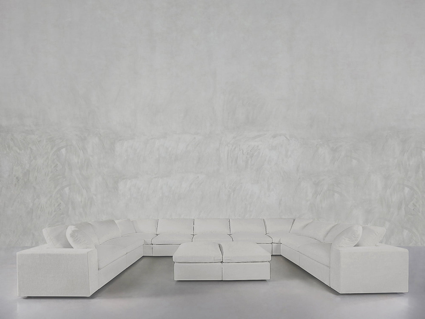 The minimalist, modern space features a 9-Seat Modular U-Sectional with Double Ottoman in white, set against a light gray textured wall and smooth gray floor, emphasizing clean lines and open space.
