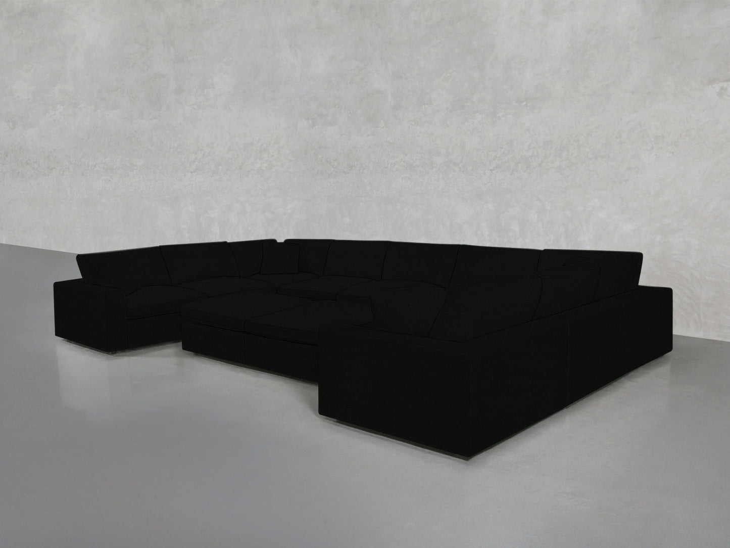 A 9-Seat Modular U-Sectional with Double Ottoman in black, featuring deep seats and clean lines, sits in an empty room with a smooth light gray concrete floor and a textured light gray wall.