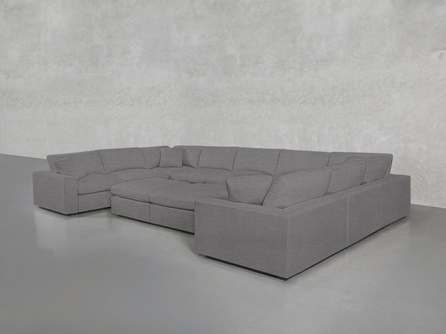 A spacious, modern 9-Seat Modular U-Sectional with Double Ottoman in gray is set in an empty room featuring a light gray textured wall and a smooth gray floor.