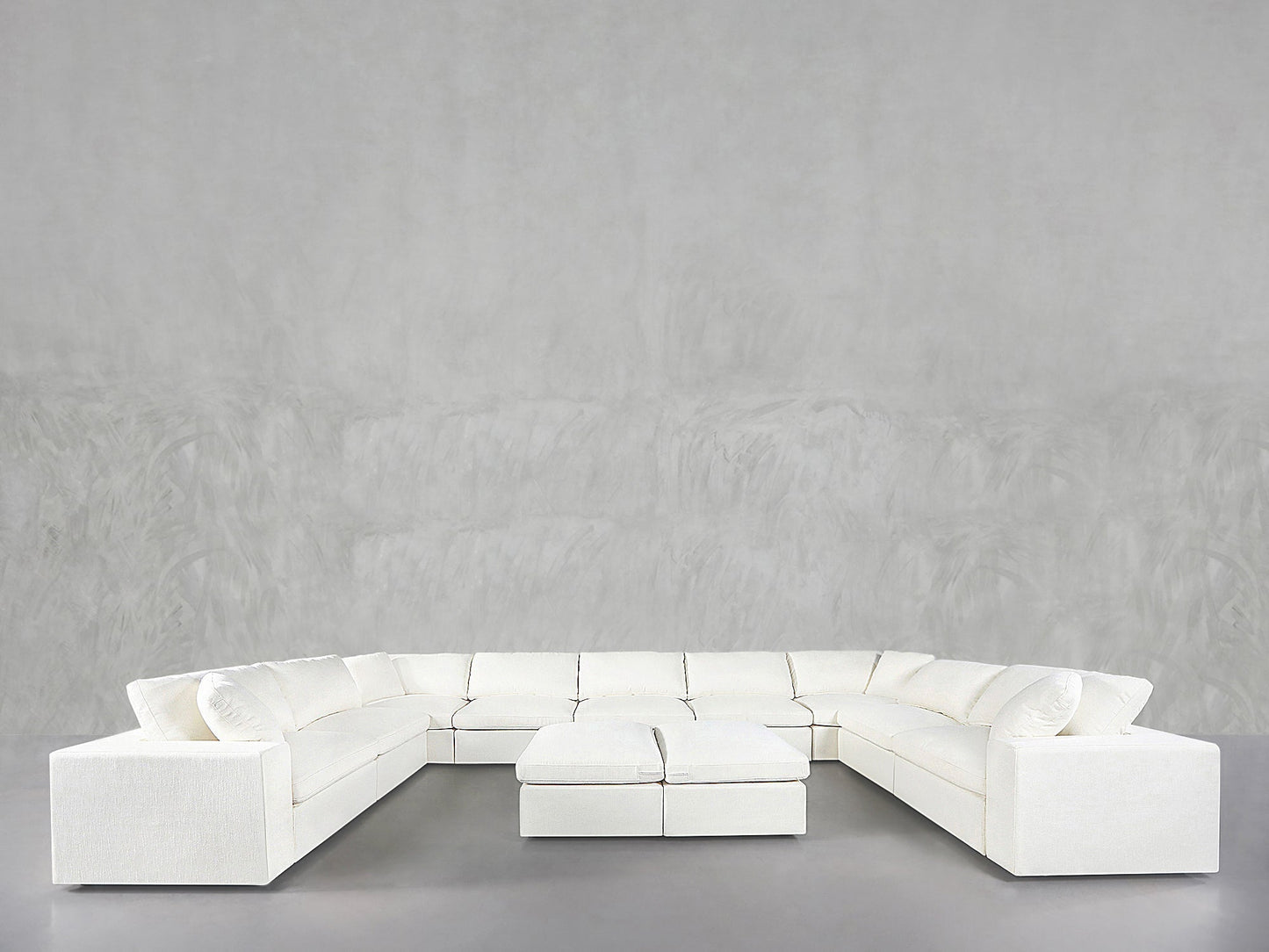 A 9-Seat Modular U-Sectional with Double Ottoman in white stands against a light gray textured wall and gray floor, creating a minimalist, modern look.
