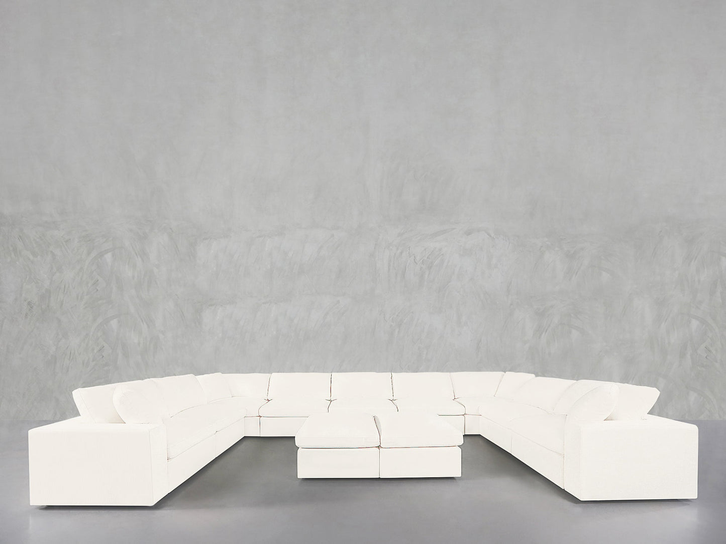 A 9-Seat Modular U-Sectional with Double Ottoman in white is arranged before a light gray, textured wall on a smooth gray floor.