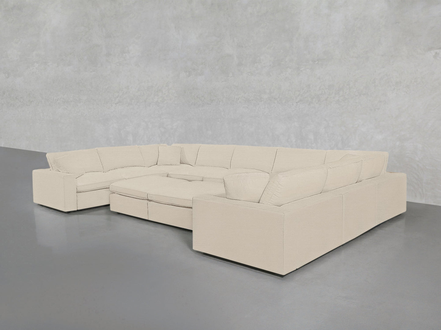 The 9-Seat Modular U-Sectional with Double Ottoman, in light beige with clean lines, thick cushions, and wide armrests, sits on a gray floor against a textured pale gray wall.