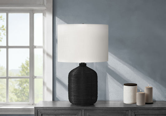 A modern rattan table lamp sits on a gray wooden console table next to three white candles, against a light blue wall near a sunlit window.