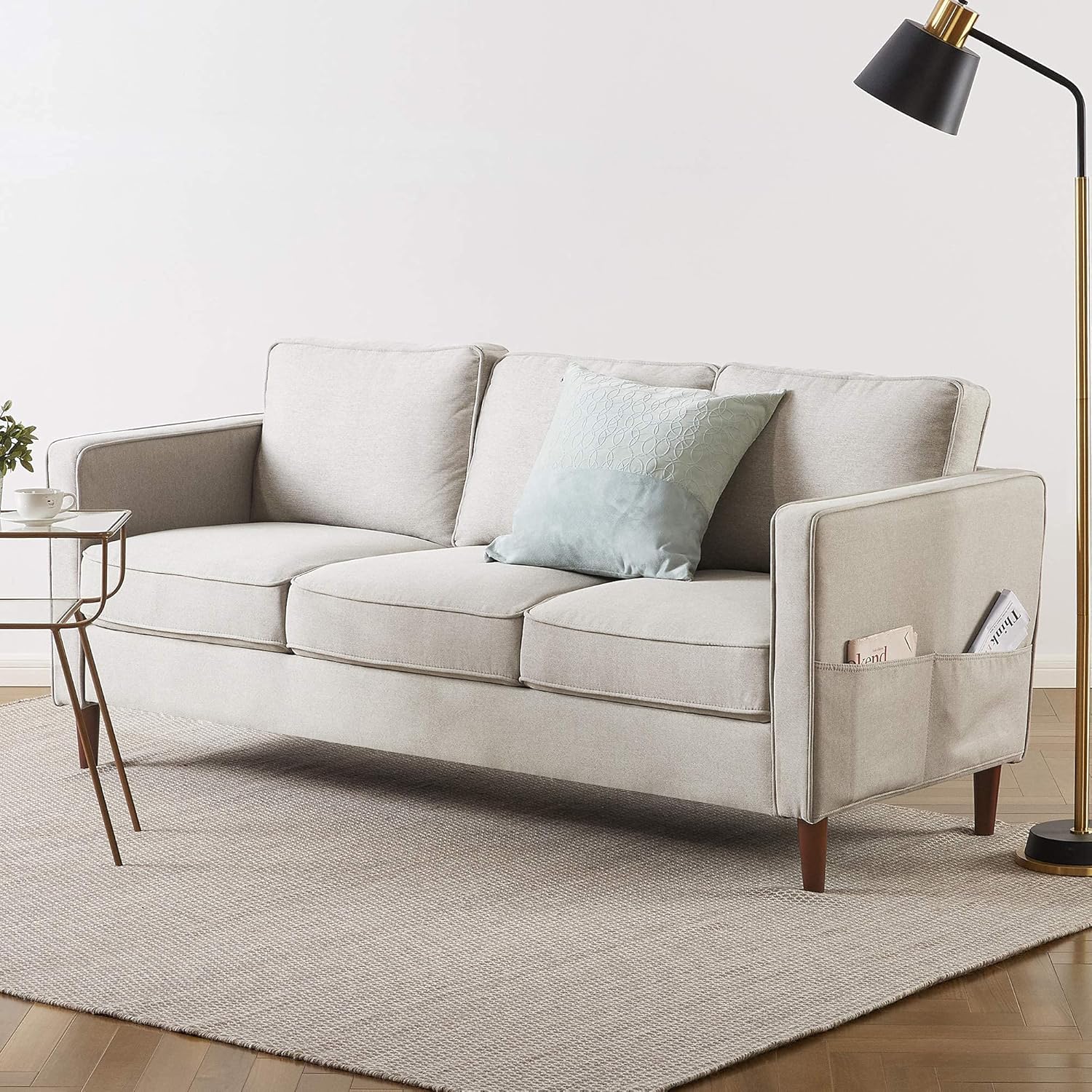 The Mellow Modern Loveseat with Armrest Pockets in light gray stands on a beige textured rug, featuring wooden legs, a light blue cushion, floor lamp to the right, side table to the left, and magazines stored in its armrest pocket.
