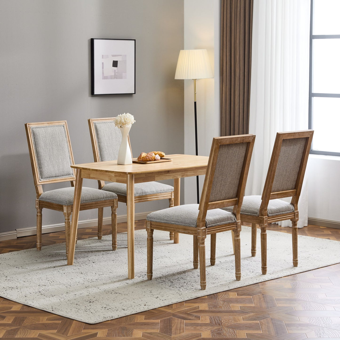 A wooden dining table with four French Country Style Dining Chairs featuring upholstered seating and fluted legs stands on a light rug. A white vase with flowers, pastries on a plate, floor lamp, framed artwork, and large window complete the room.