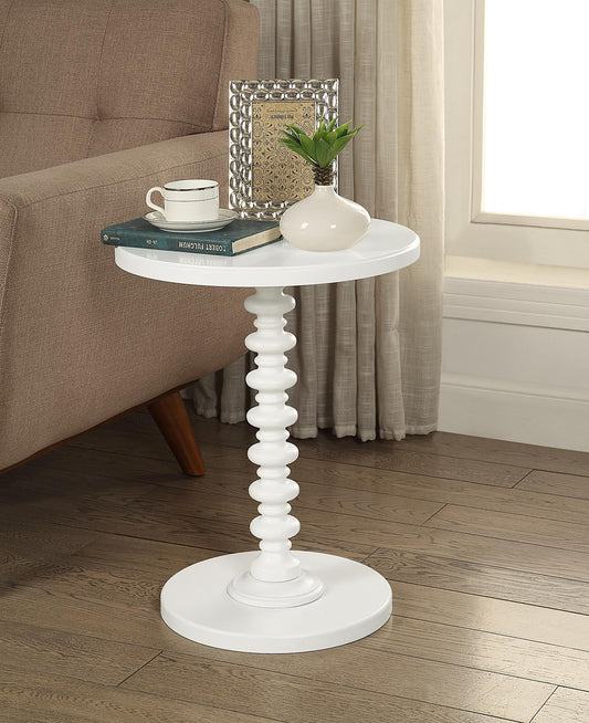 The Acton - Accent Table - White, featuring a round top and sculpted pedestal base, stands on a wood floor beside a beige sofa. On it are two books, a white mug, a framed photo, and a small white vase with green leaves.