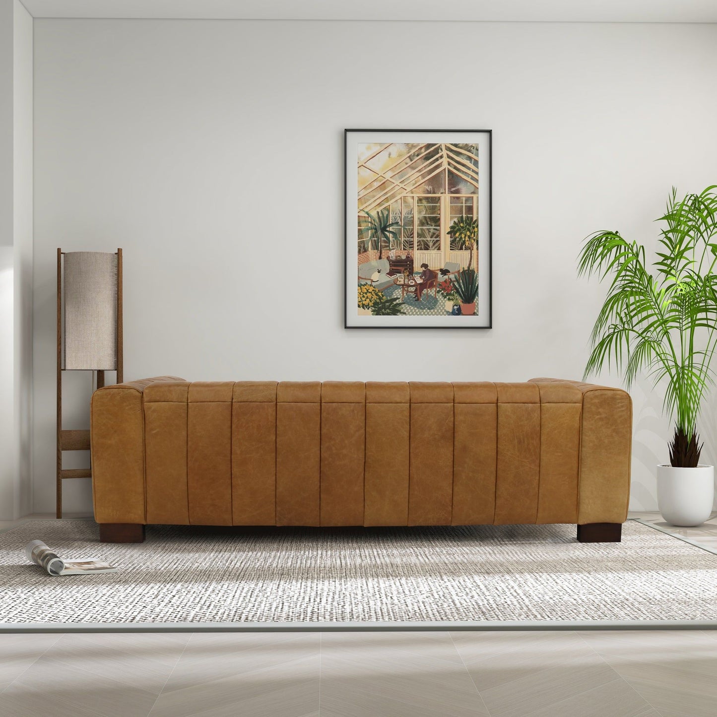 A modern living room featuring the Dixon - Waxy Sofa in tan, a tall floor lamp, a rolled-up magazine on the floor, a potted palm plant, and framed botanical art on a white wall.