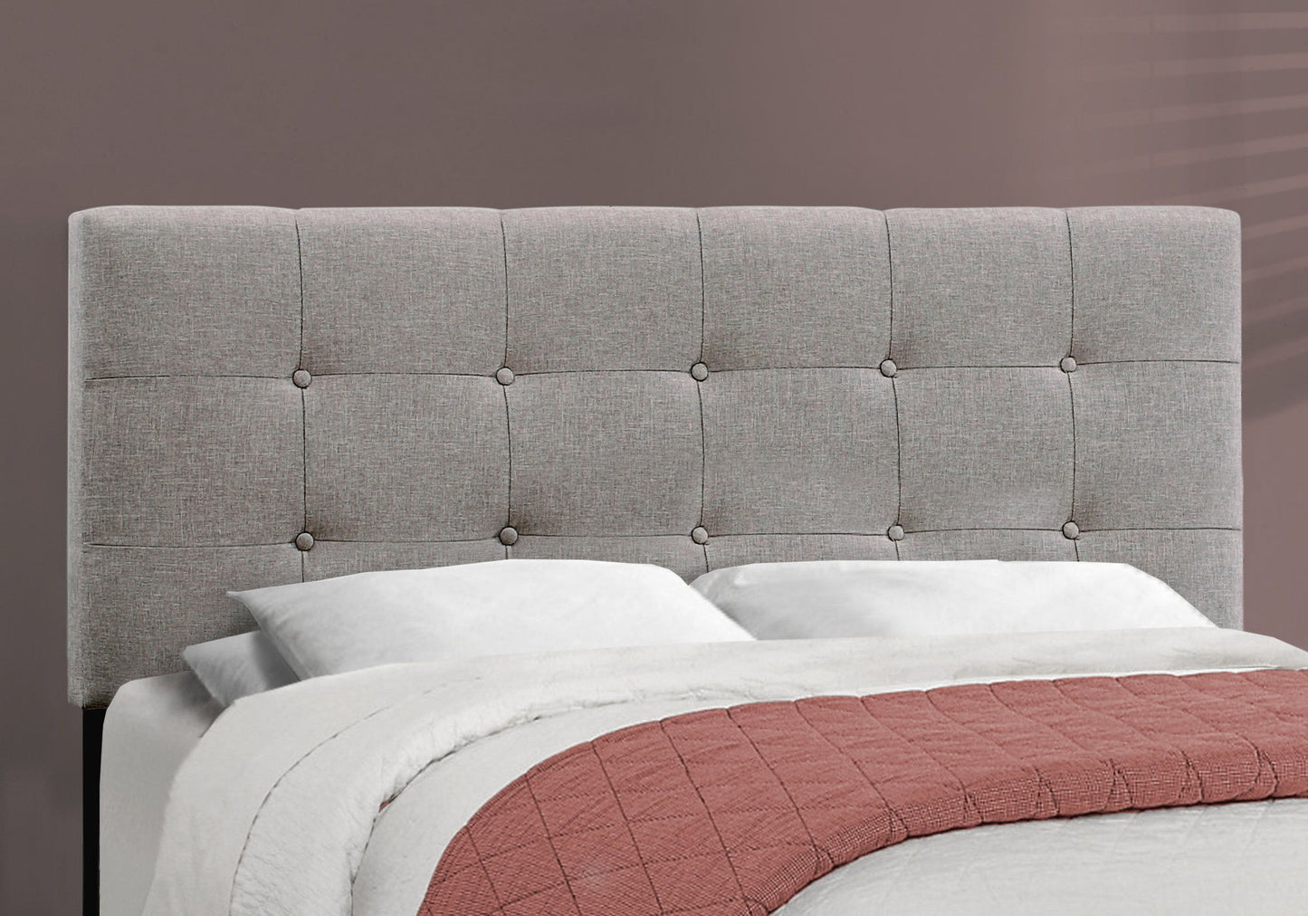 A transitional upholstered bed frame with a light gray tufted headboard, white pillows and bedding, a mauve quilt at the foot, all set against a taupe wall.