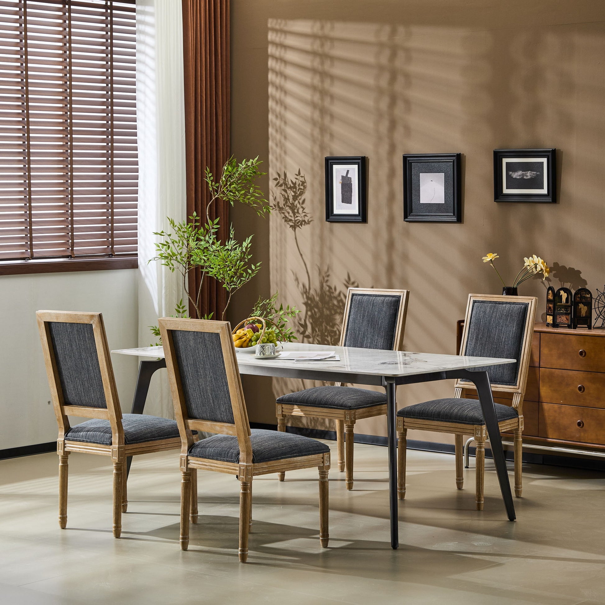 A modern dining room features a rectangular white table with four French Country Style Dining Chairs with upholstered seating and fluted legs, a corner plant, framed wall art, and a sideboard lit by soft natural light from window blinds.