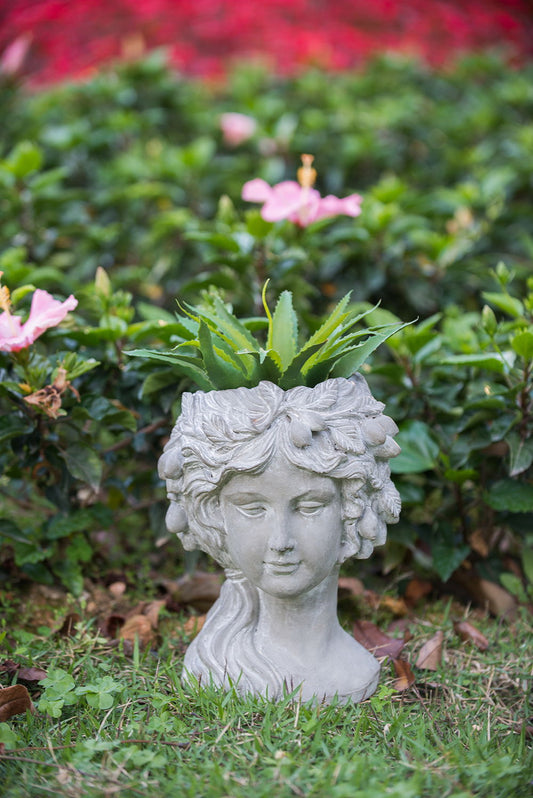 The Cement Bust Wall Planter Indoor Outdoor Home Garden Decor, 8 - Gray, features a woman's head design with plants growing from the top, shown on grass in a garden with pink flowers and lush green foliage in the background.