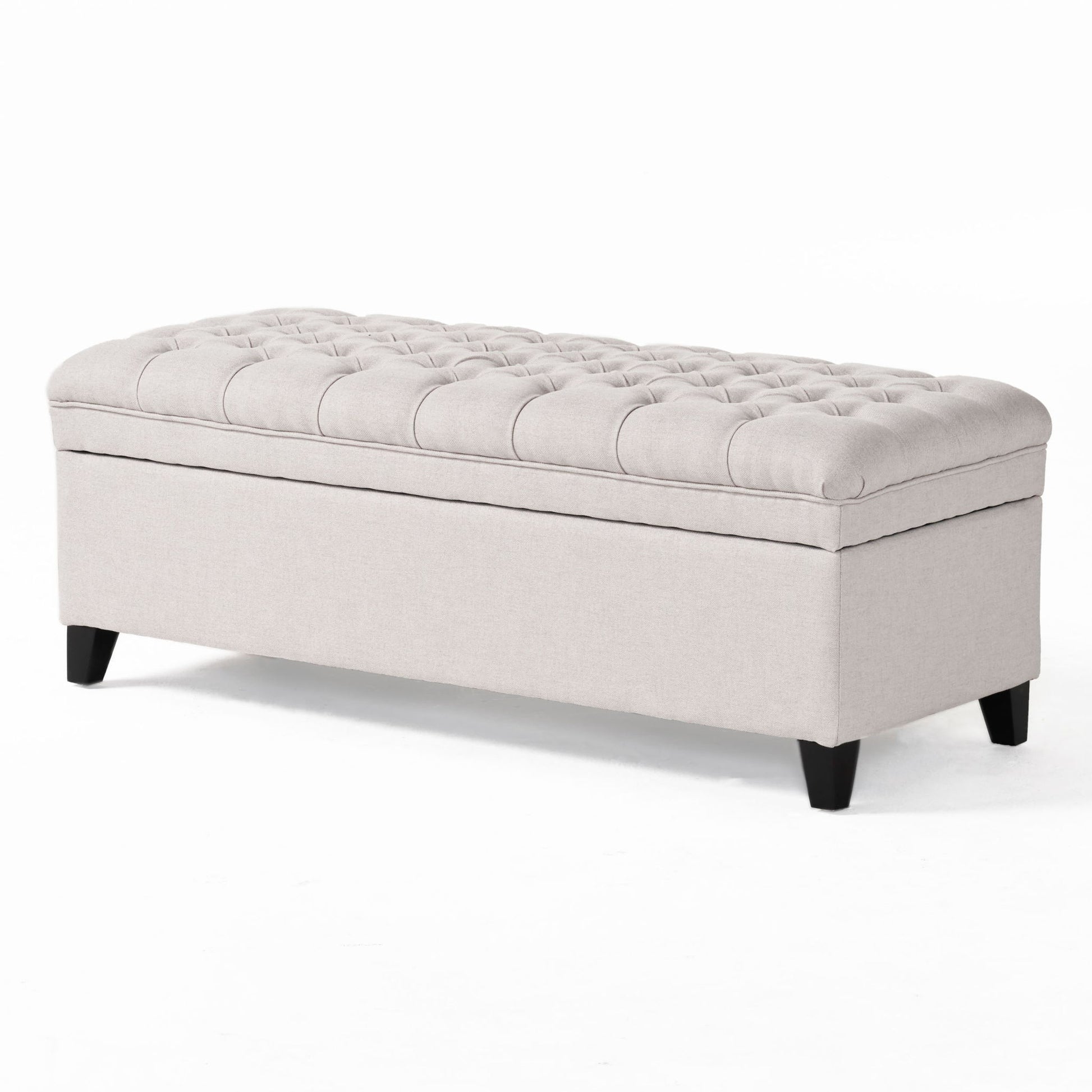 The Juliana Storage Ottoman features a rectangular, light beige upholstered design with a tufted cushion top and short black birch legs, shown on a white background.