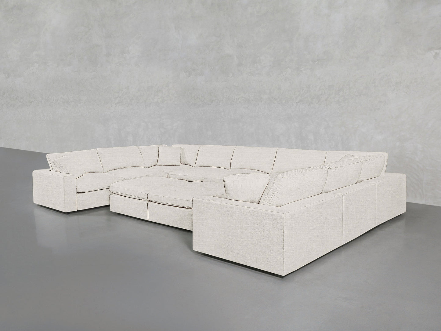 The 9-Seat Modular U-Sectional with Double Ottoman in light beige features wide armrests and plush cushions, arranged on a smooth gray floor against a textured pale gray wall.