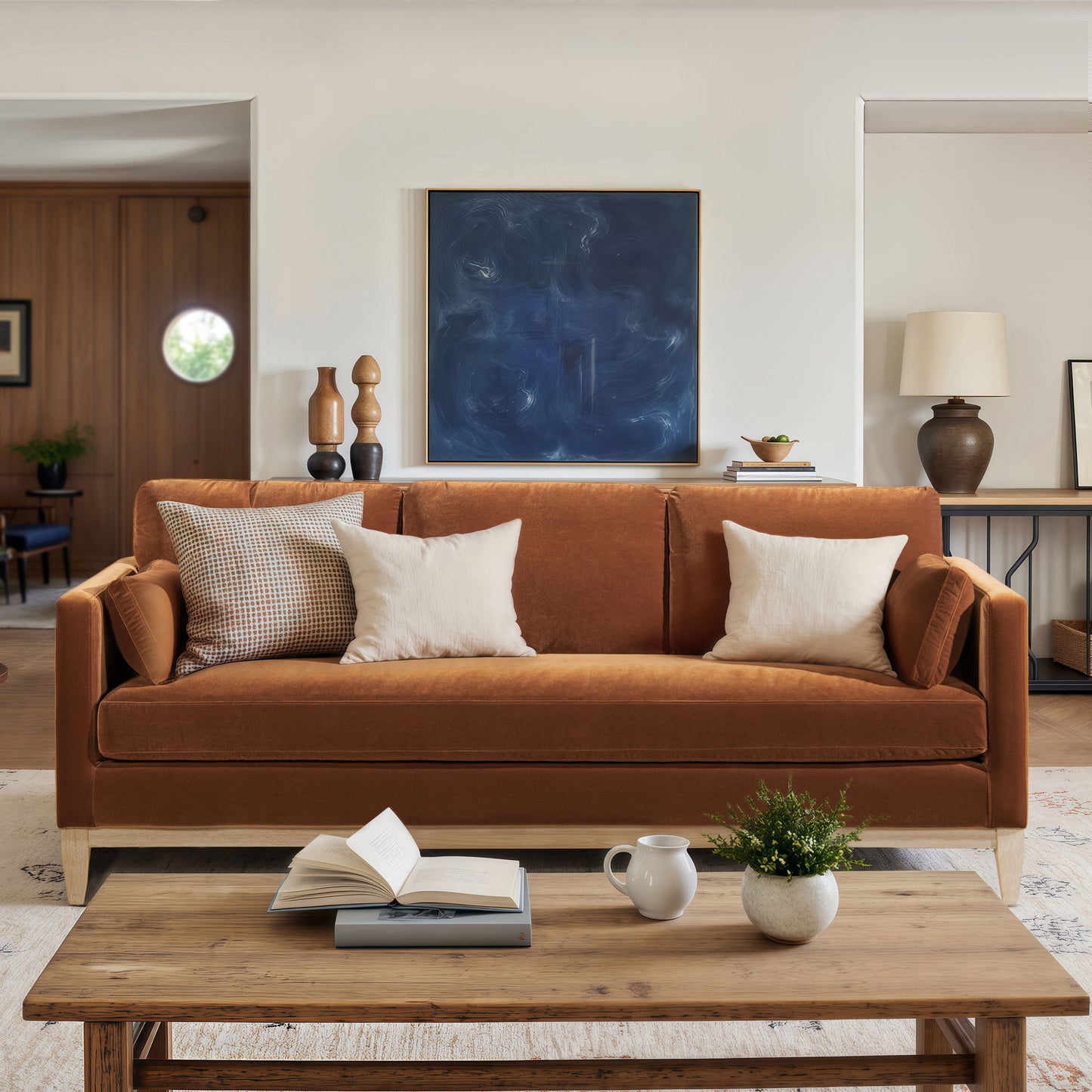 A modern living room featuring the Knox - Modern Farmhouse Sofa in brown velvet with three white and beige pillows, a wooden coffee table with books, a plant, ceramic set, a dark abstract painting, and neutral decor.