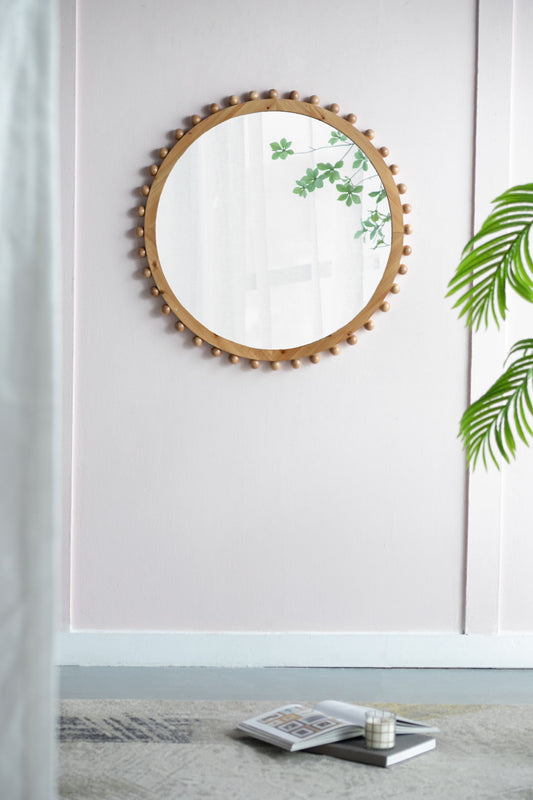 The Wood Round Mirror With Beaded Frame in brown hangs on a light pink wall. To the right, a leafy green plant is visible, while a stack of books and a candle rest on the light-colored carpet below.
