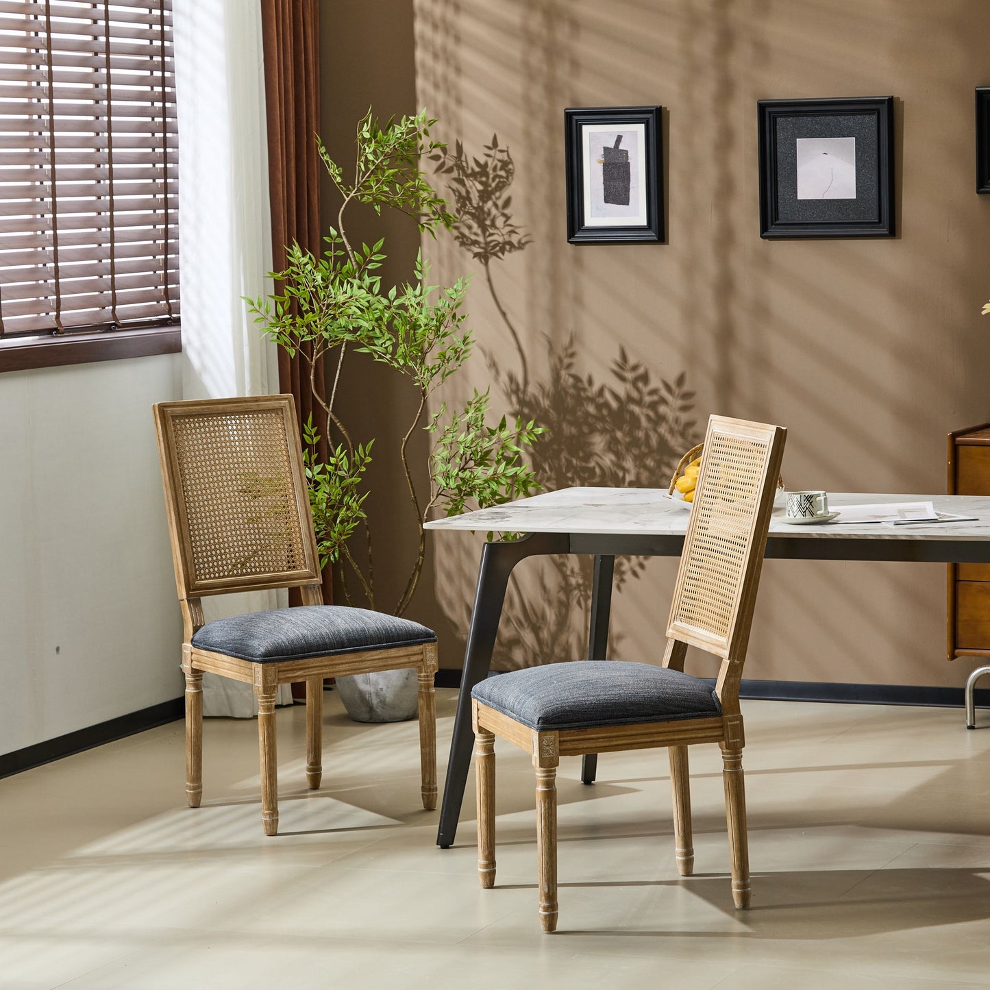 Two French Country Dining Chairs with rectangular cane backrests and smooth gray upholstered seats sit by a white table in a sunlit room with beige walls, green potted plant, wall art, and window blinds casting patterned shadows.