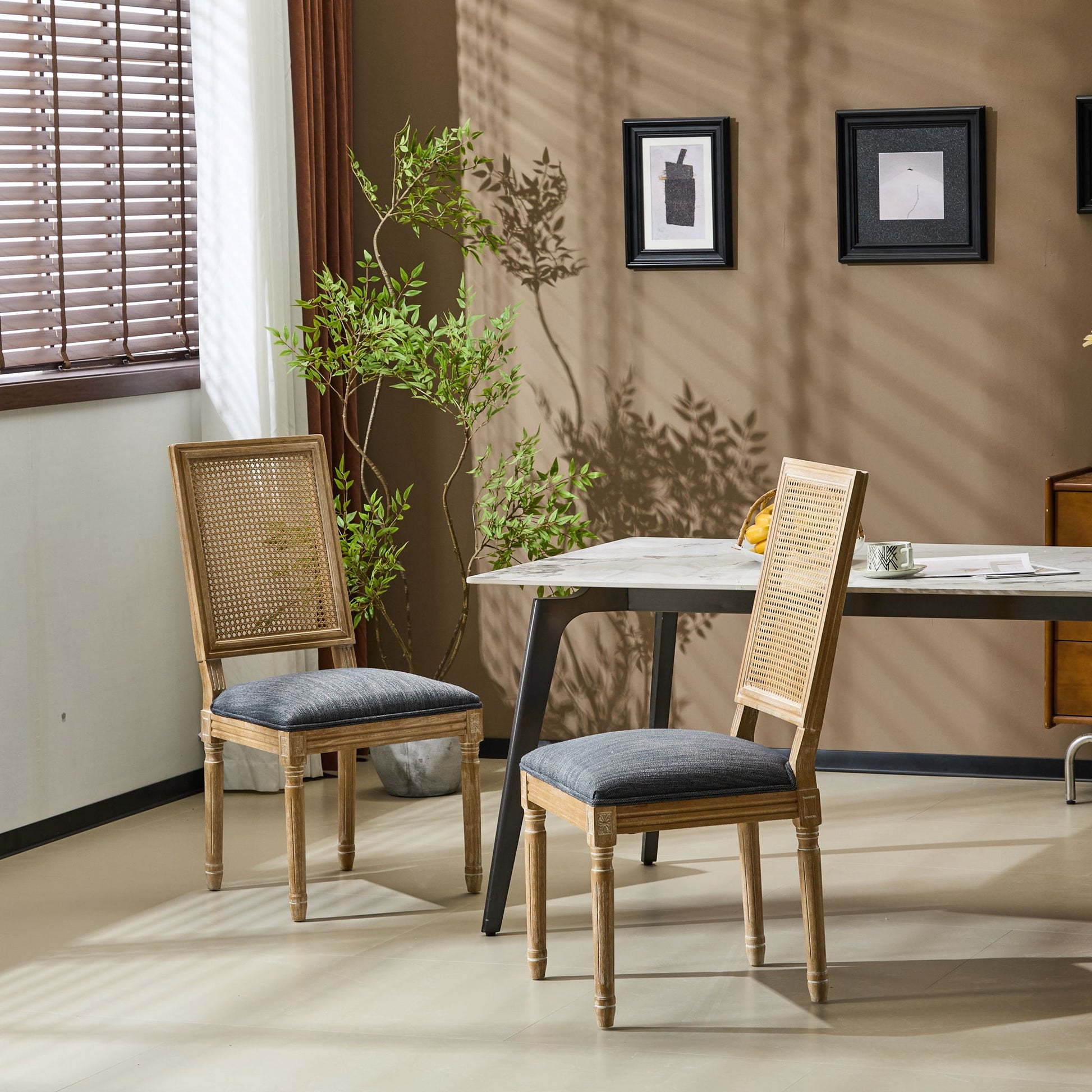 Two French Country Dining Chairs with rectangular cane backrests and smooth gray upholstered seats sit by a white table in a sunlit room with beige walls, green potted plant, wall art, and window blinds casting patterned shadows.