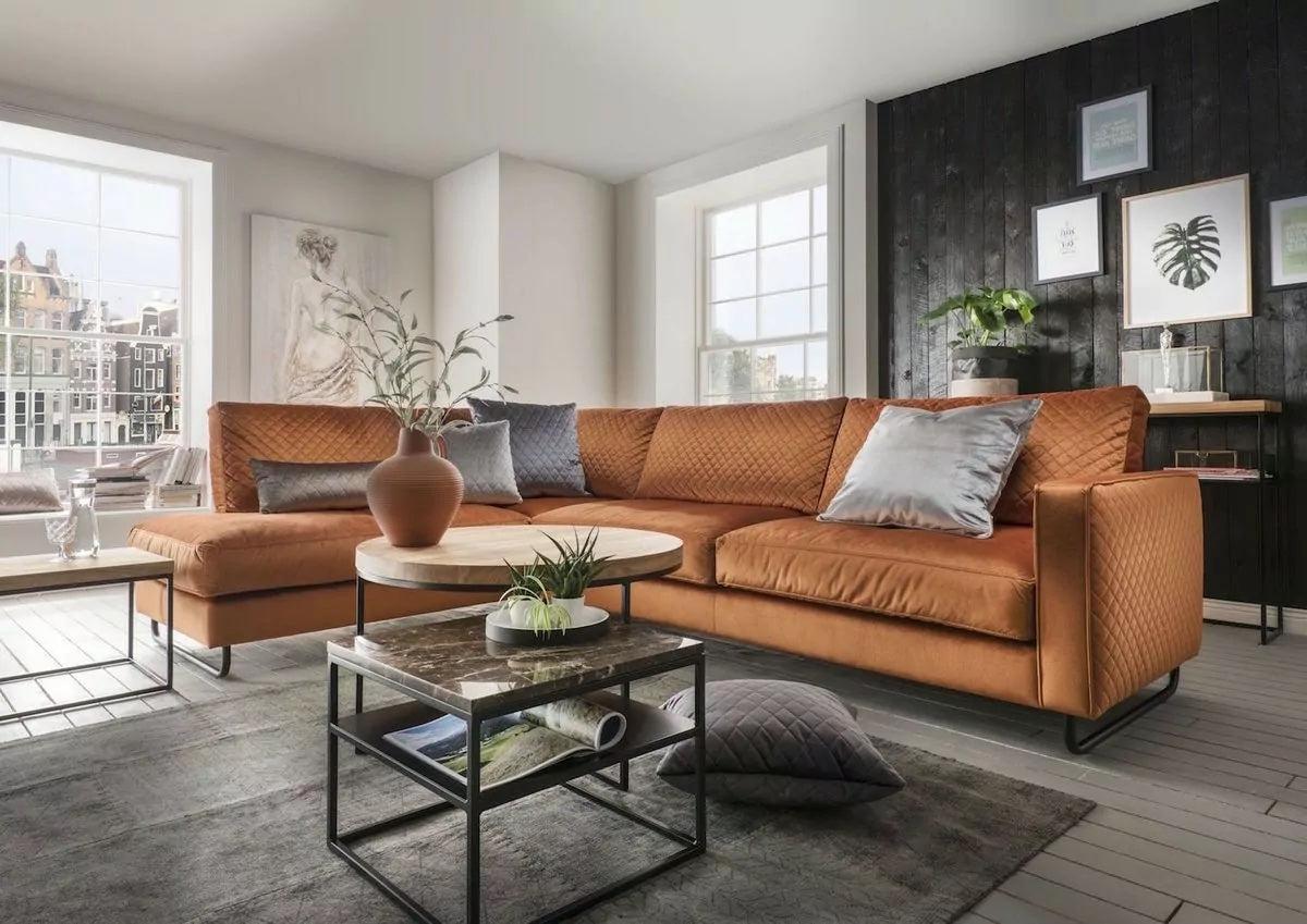 A cozy living room features a large orange sectional sofa with several cushions, a round coffee table with a potted plant and books, and a dark accent wall adorned with framed artwork. Large windows let in ample natural light, illuminating the space.