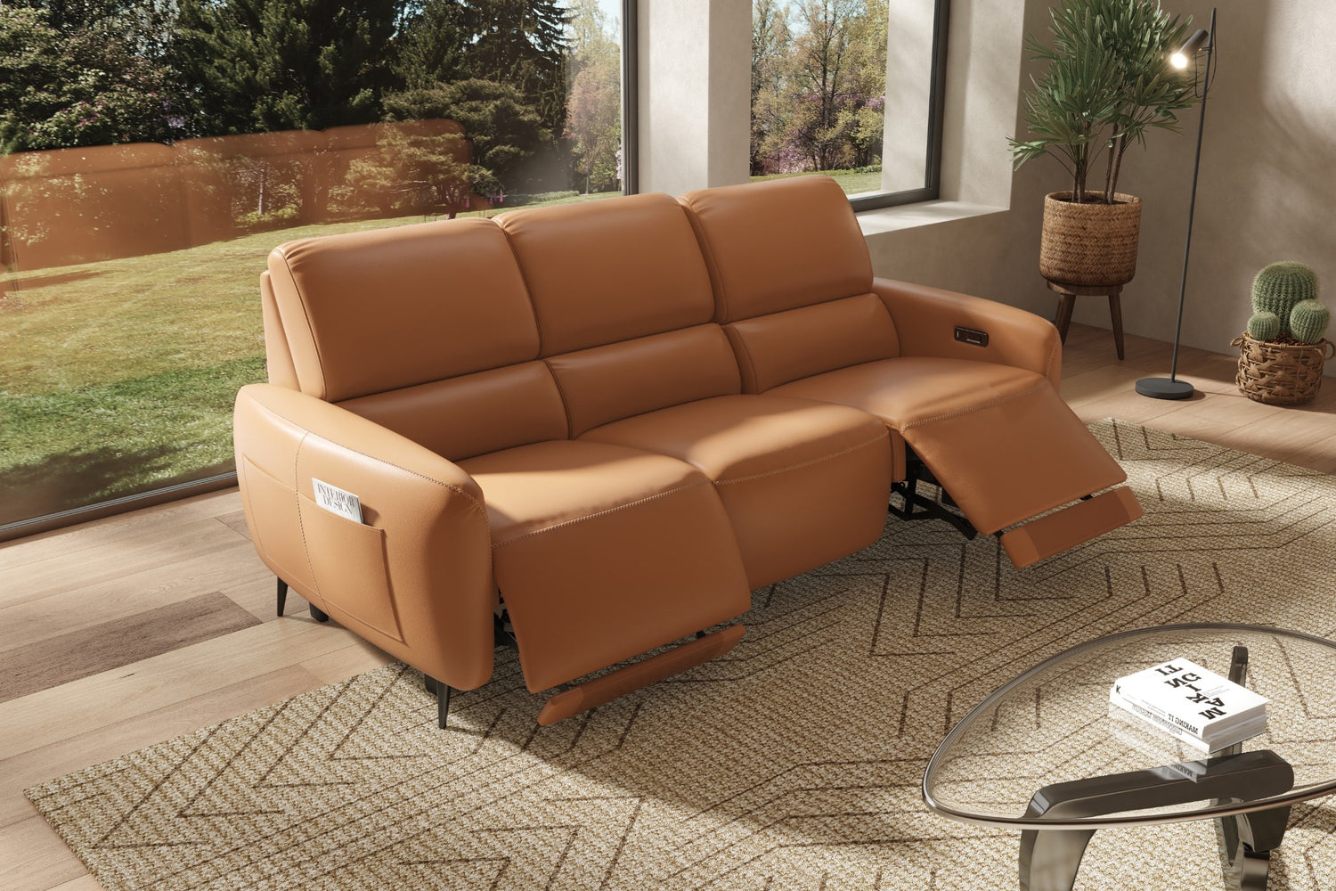 A modern living room with a tan leather recliner sofa, partially extended. A light rug is on the wooden floor, and a glass coffee table holds a couple of magazines. Large windows reveal greenery outside. Potted plants and a woven basket decorate the space.
