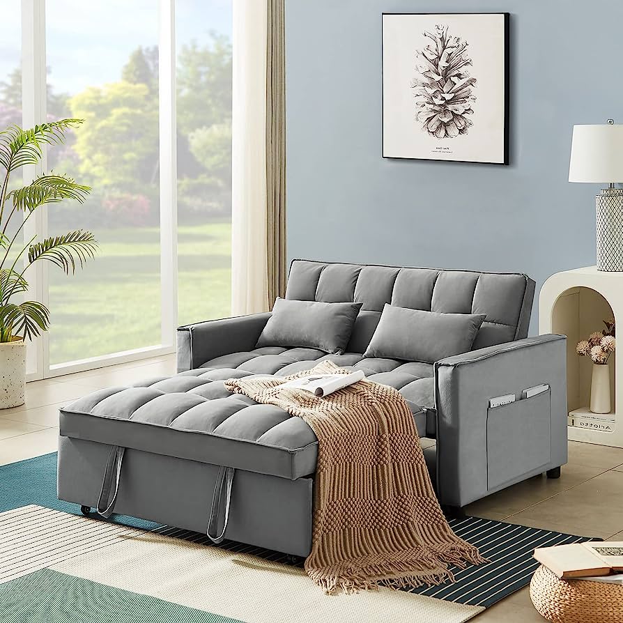 A cozy living room featuring a gray tufted sofa bed with matching throw pillows. A beige and brown striped blanket is draped over the ottoman, which is attached to the sofa. The room has a large window with greenery outside, a potted plant, and a side table with a lamp and decor. A framed pinecone artwork hangs on the blue wall.
