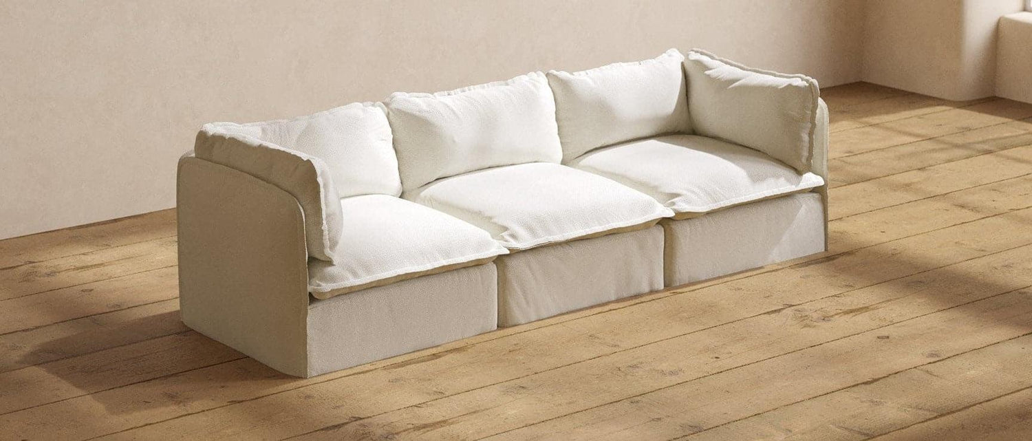 A beige, three-seater sofa with plush white cushions is placed against a neutral beige wall on a wooden floor. The room is well-lit, casting soft shadows on the floor and highlighting the sofa's comfortable and cozy appearance.