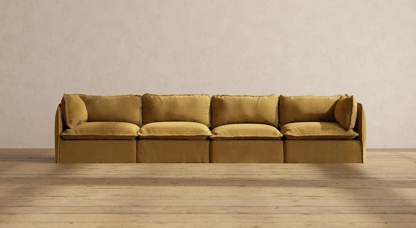 A mustard yellow, modern sectional sofa with four cushioned seats and matching backrest pillows is centered against a plain, light beige wall. The sofa sits on a smooth wooden floor, enhancing the minimalist aesthetic of the room.