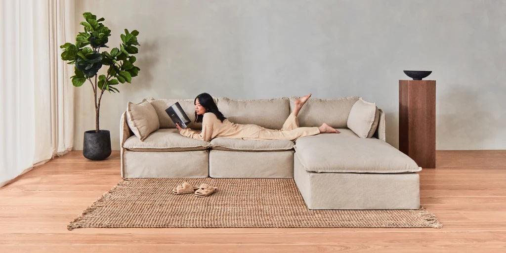 A woman in comfortable clothing lounges on a large beige sectional sofa, reading a book. A jute rug, a pair of slippers, a tall indoor plant, and a wooden pedestal with a bowl on it complete the minimalist living room setting.
