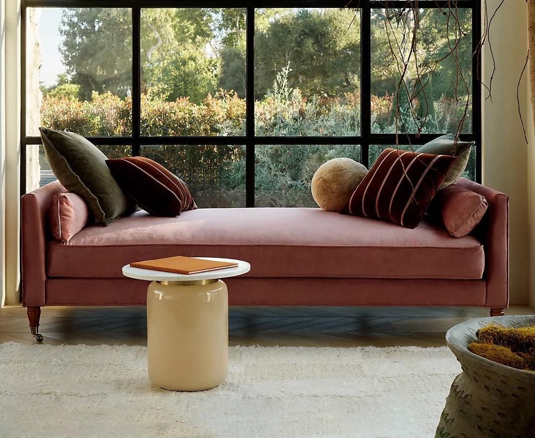 A cozy room features a plush pink daybed with wooden legs, adorned with gray and rust-colored pillows, beside a large window showcasing lush greenery outside. A white cylindrical side table with a notebook on it is placed in front of the daybed on a white rug.