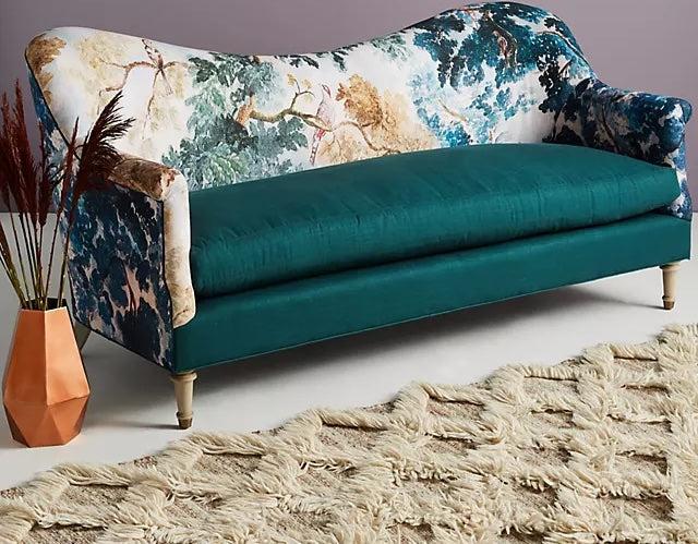 A teal blue and white upholstered sofa with an artistic tree-patterned backrest stands on a light grey floor. In front of it lies a shaggy beige rug with a geometric pattern. To the side, there is an orange geometric vase with dried flowers.