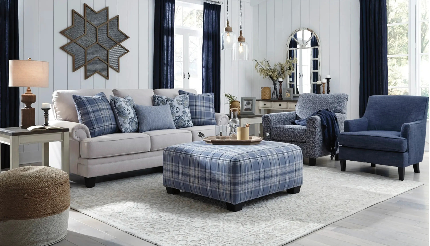 A stylish living room with a light beige sofa adorned with blue plaid and patterned pillows, a large plaid ottoman, a blue upholstered armchair, and a wooden coffee table. The room features dark blue curtains, wall art, a large mirror, and decorative lighting.