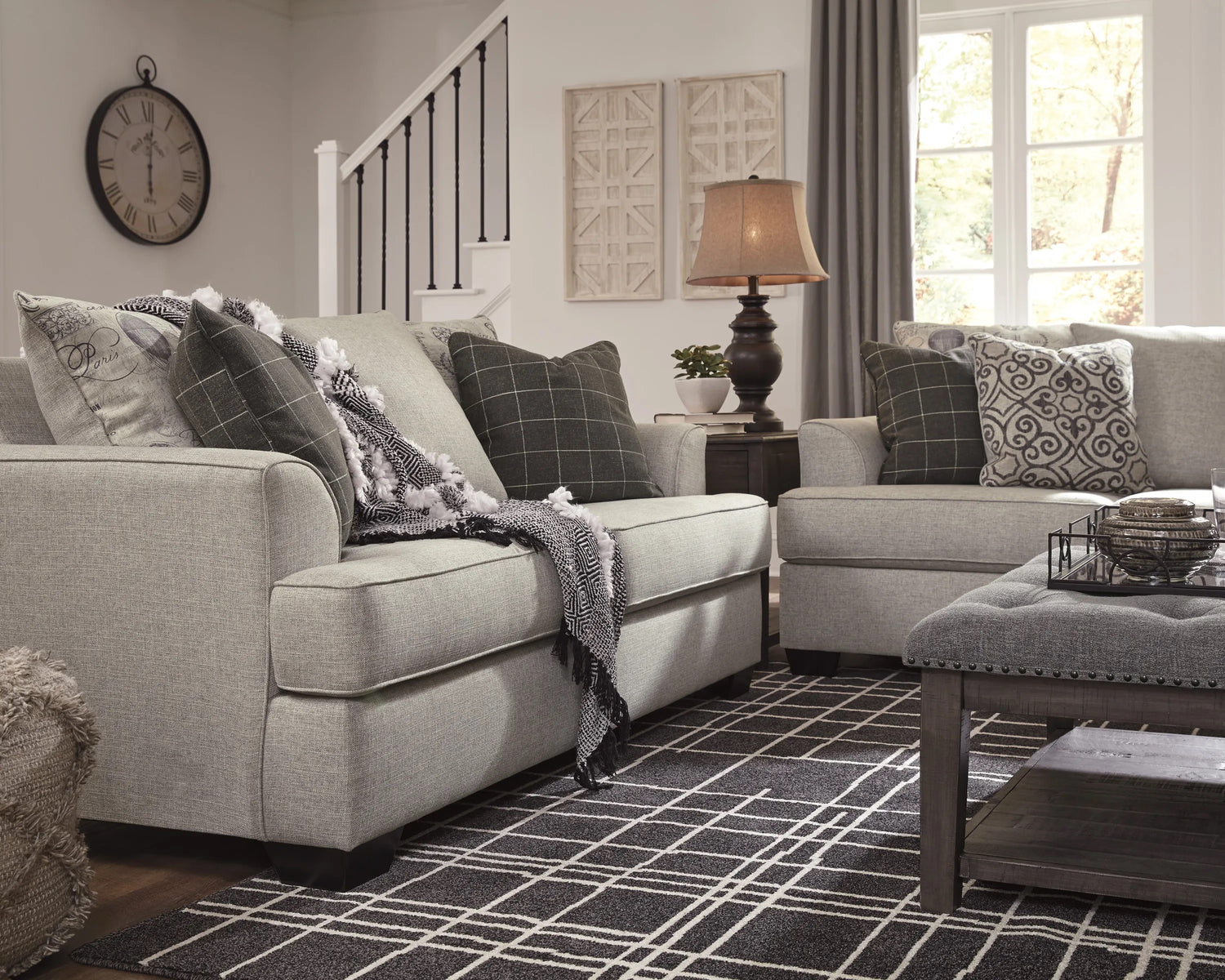 A cozy living room with two gray sofas adorned with patterned pillows and a monochrome throw blanket. A coffee table with a decorative tray sits on a black-and-white geometric rug. A lamp on a side table, abstract wall art, and a large clock complete the decor.