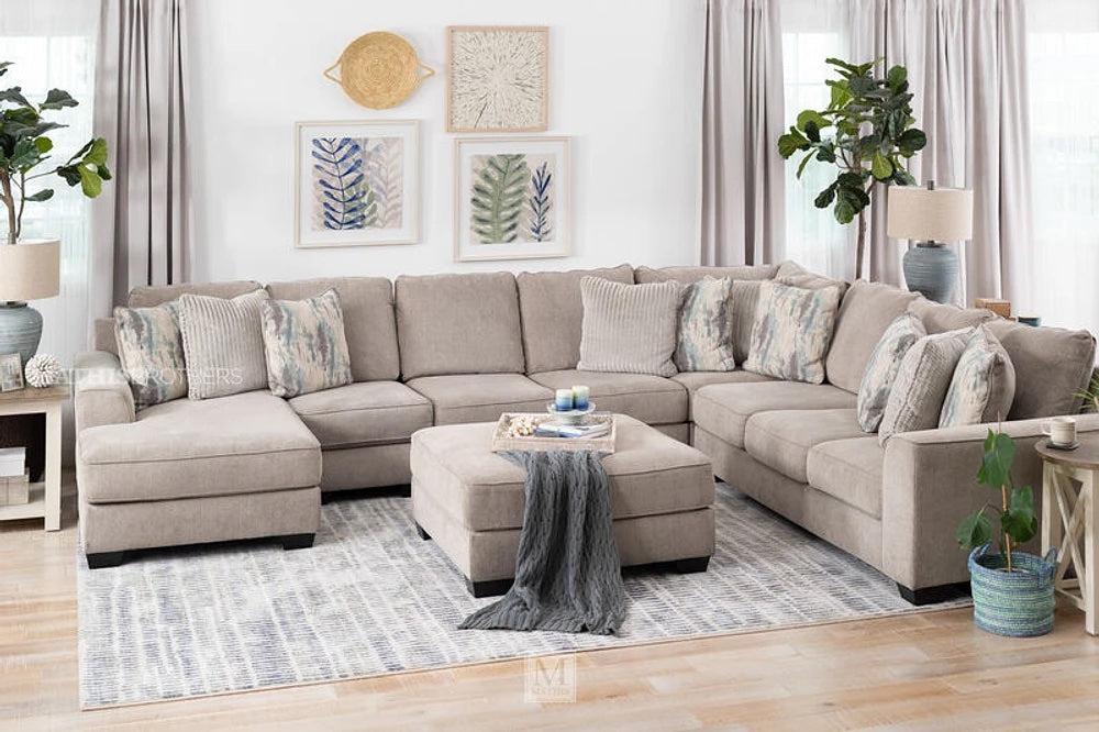 A spacious living room features a large L-shaped beige sectional sofa adorned with patterned and solid pillows. The space is complemented by a matching ottoman, coastal-themed wall art, green plants, and neutral curtains. Natural light fills the room, highlighting a light grey rug.