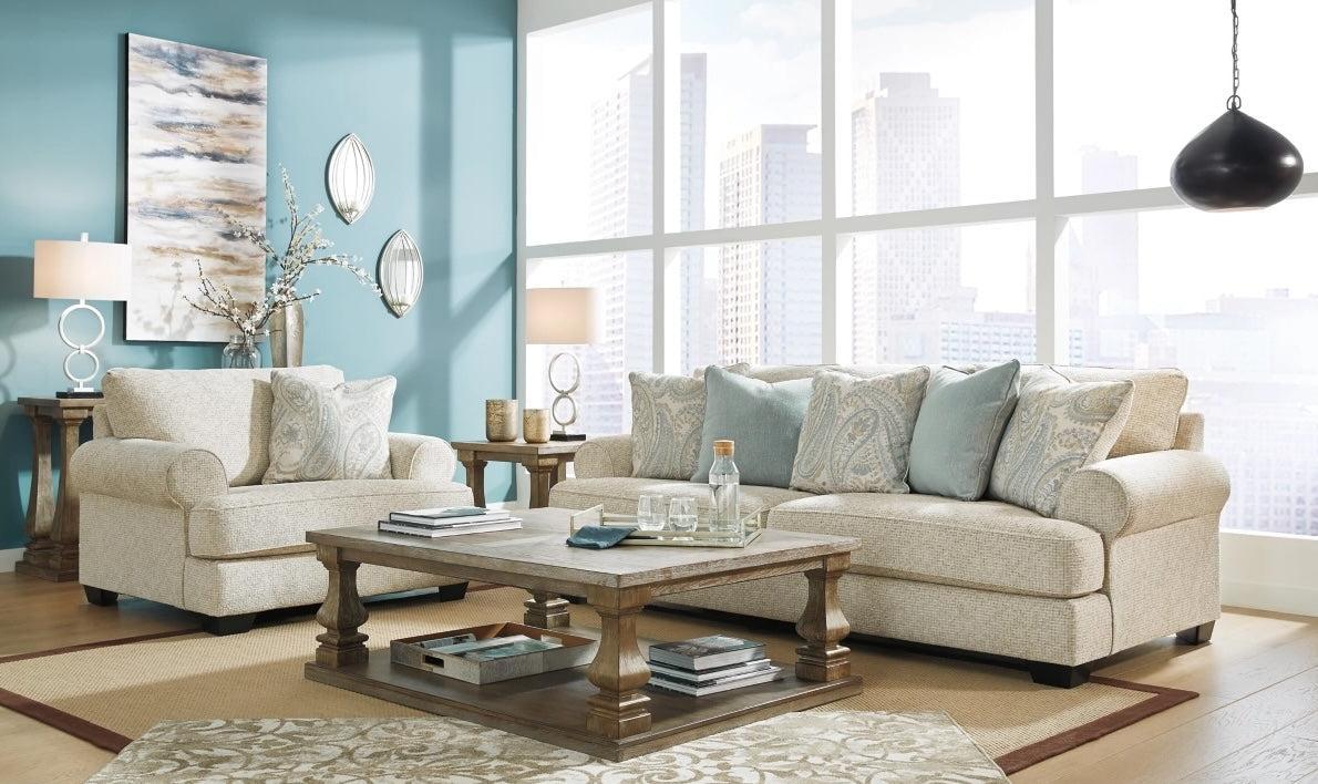 A modern living room with light beige sofas adorned with pastel blue and patterned pillows. A wooden coffee table sits in the center on an area rug. Large windows offer a cityscape view. The walls are painted teal and decorated with abstract artwork and mirrors.