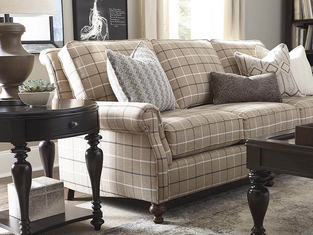 A cozy living room with a beige plaid sofa adorned with various patterned pillows. A round, dark wood side table with a lamp and decorative plant sits to the left of the sofa. A rectangular dark wood coffee table is in front, and framed art is on the wall.
