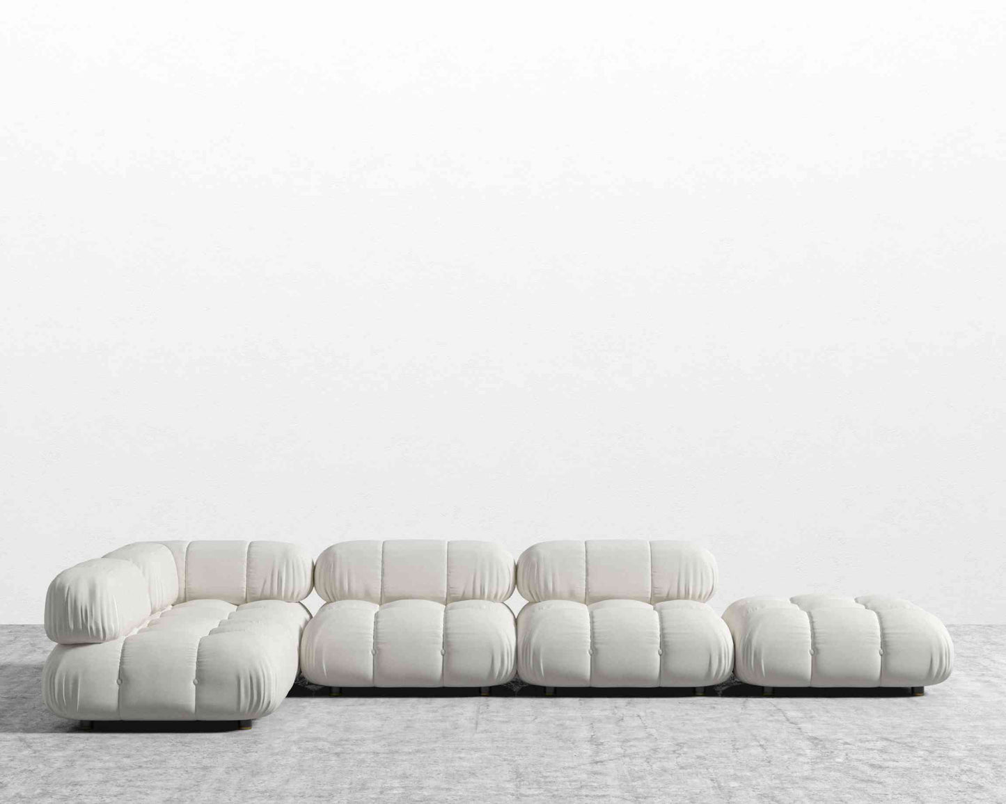 The Belia Modular Sectional, featuring white, tufted cushions, is arranged in an L-shape against a plain white wall on the gray carpeted floor. This sofa design showcases low-profile seating and a minimalist aesthetic.