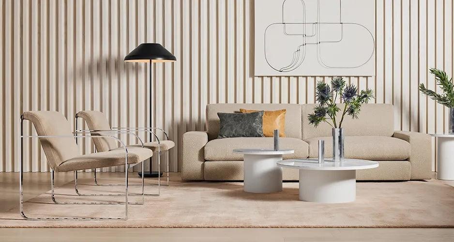 A modern living room featuring a beige sofa with two cushions, two beige chairs with metal frames, and two round white coffee tables with vases. A black floor lamp stands in the corner, and abstract wall art hangs above the sofa. The wall has vertical wooden panels.
