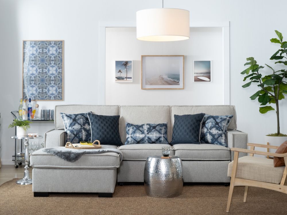 A modern living room with a light grey sofa adorned with blue-patterned cushions. Above the sofa are three framed landscape photos. There is a round metallic side table, a wooden chair, a hanging white lamp, and a potted plant in the background.