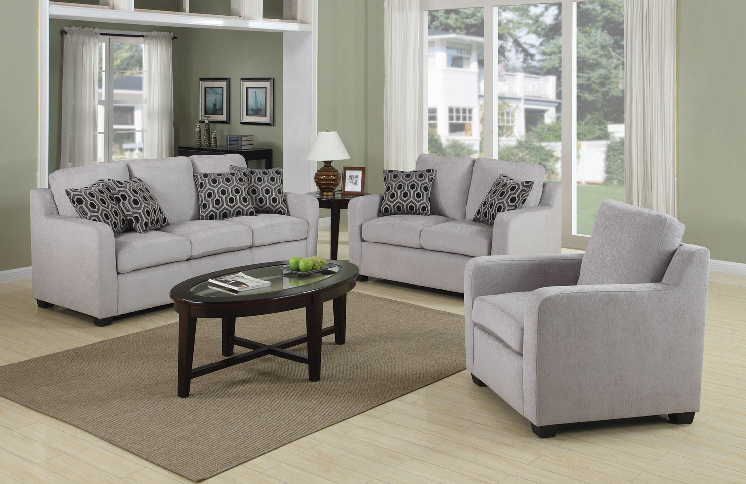 A modern living room with two gray sofas and a matching armchair, all adorned with patterned black and white throw pillows. A wooden oval coffee table sits on a beige rug in the center, with a lamp and decorative items. Large windows provide ample natural light.