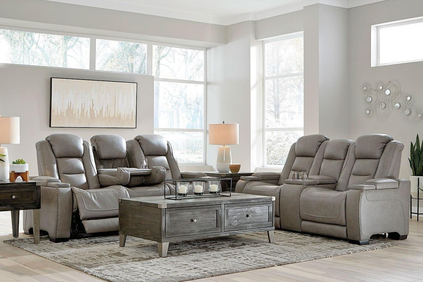 A modern living room features two gray leather reclining sofas, a wooden coffee table with storage drawers, and two side tables with lamps. The space is adorned with a wall painting, decorative wall art, and large windows that let in natural light.