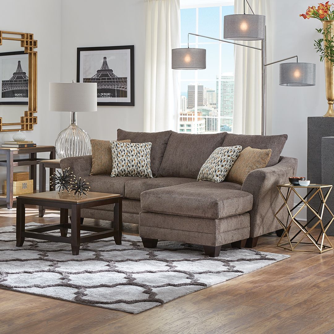 A modern living room featuring a brown sectional sofa with patterned pillows. A large floor lamp with multiple shades stands behind the sofa. A coffee table with a decorative piece and a side table with a lamp are also present. The room has framed artwork and a patterned rug.