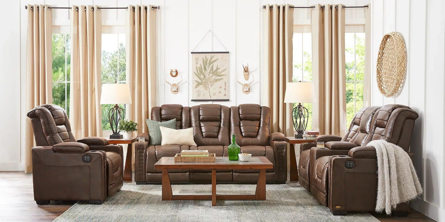 A cozy living room features three brown leather recliners arranged around a wooden coffee table. The space is accented by beige curtains, two lamps, wall art with botanical prints, and a woven wall hanging. The decor includes books and green glass bottles on the table.